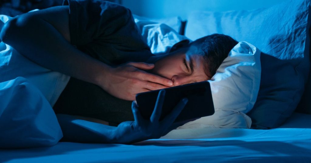 Man looking at his mobile phone in the middle of the night because he is suffering from sleep deprivation. 
